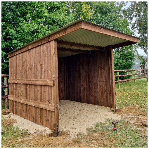 Abri pour chevaux en madrier, vue de profil droit, avec une barrière à 3 lisses en arrière-plan.